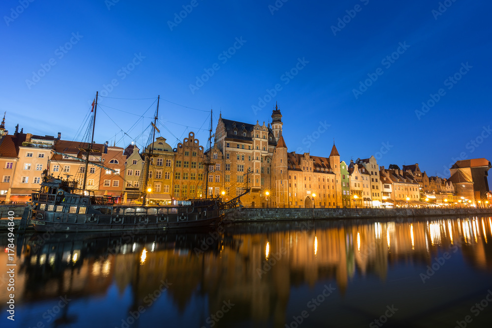 Scenic view of a tourist sailing boat and lit old buildings along the Long Bridge waterfront at the Main Town in Gdansk, Poland, in the evening.