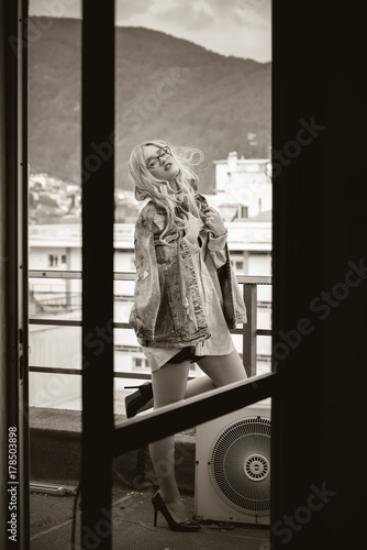 Blonde woman standing on a balcony wearing shorts, jean jacket and high heels, cityscape as background