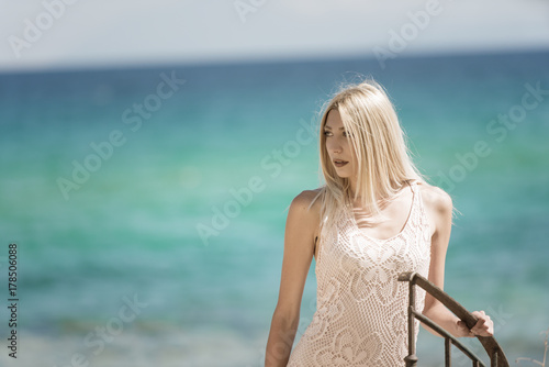 Blonde woman in vintage knit dress with crochet patterns standing on stairs, ocean as background © Domforstock