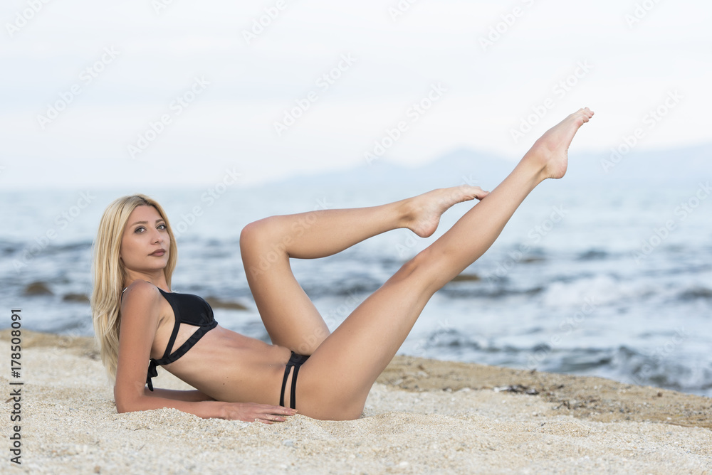 Slim tall gorgeous blonde woman at the beach lying on sand lifting her long legs
