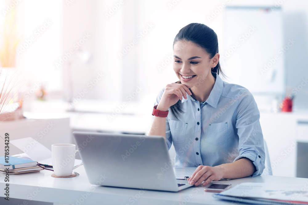 Beaming young lady looking at laptop and smiling at work Beaming young lady looking at laptop and smiling at work