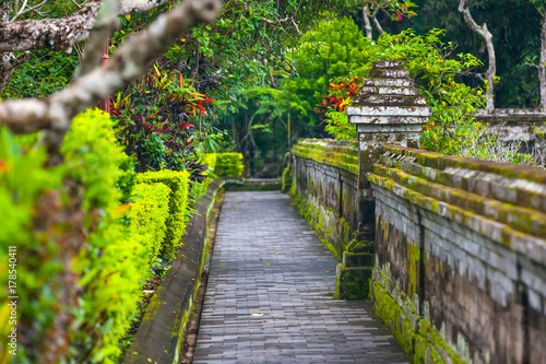 The path around the temple complex is surrounded by green tropical trees and cut bushes along the stone wall with moss. Taman Ayun Temple of Mengwi Empire  Badung regency  Bali  Indonesia.