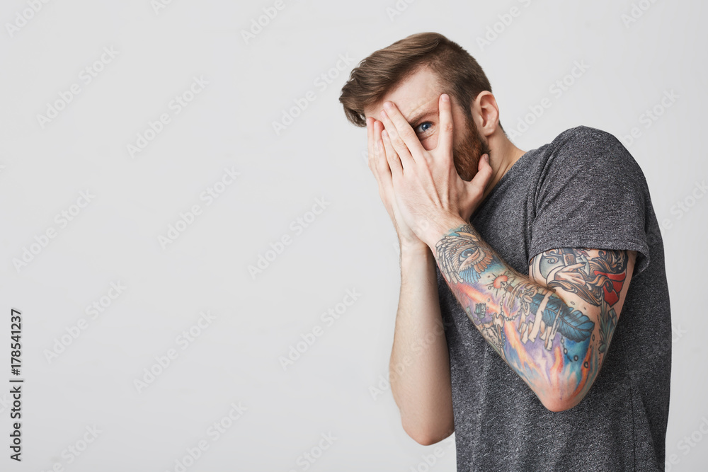 Close up portrait of young good-looking bearded tattoed guy with fashionable short haircut in casual gray t-shirt closing face with hands, looking through fingers with one eye can't look at violent Close up portrait of young good-looking bearded tattoed guy with fashionable short haircut in casual gray t-shirt closing face with hands, looking through fingers with one eye can't look at violent