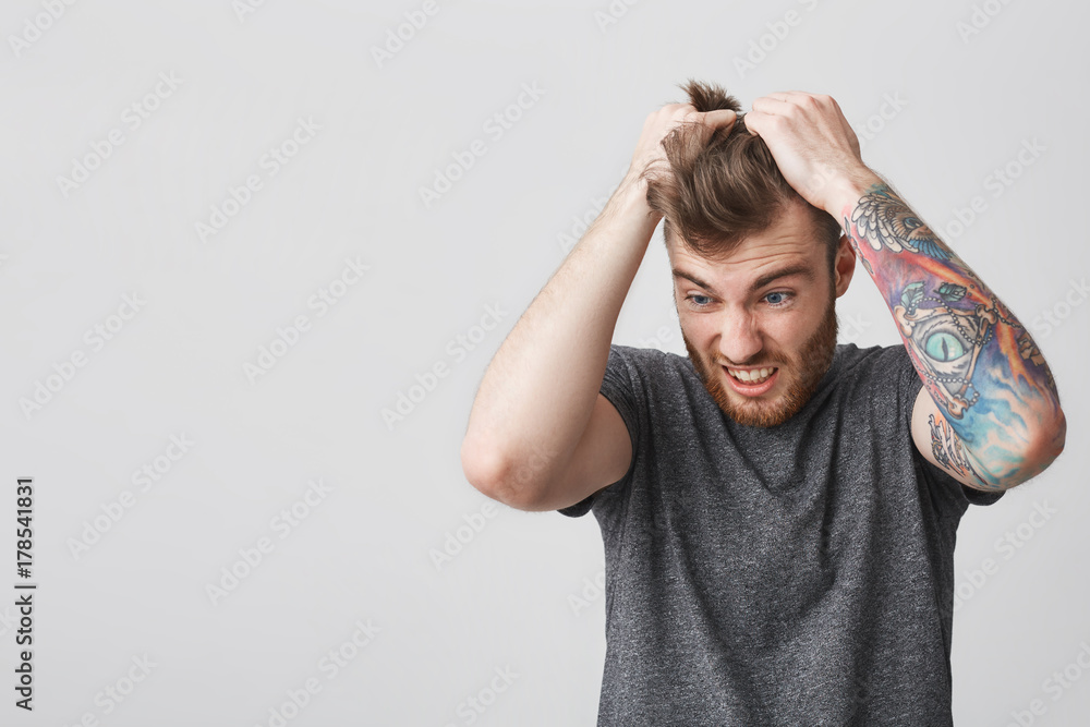Portrait of stressed young good-looking bearded man with stylish hairstyle  and tattoo on left arm in casual gray shirt squizing hair with hands,  looking aside with frustrated and angry face expression Stock