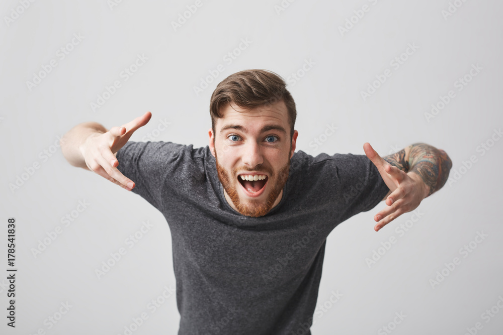 Close up of young cheerful bearded tattooed good-looking guy with stylish hairstyle in casual gray shirt spreading hands, lens in camera with happy and enthusiastic expression. Close up of young cheerful bearded tattooed good-looking guy with stylish hairstyle in casual gray shirt spreading hands, lens in camera with happy and enthusiastic expression.