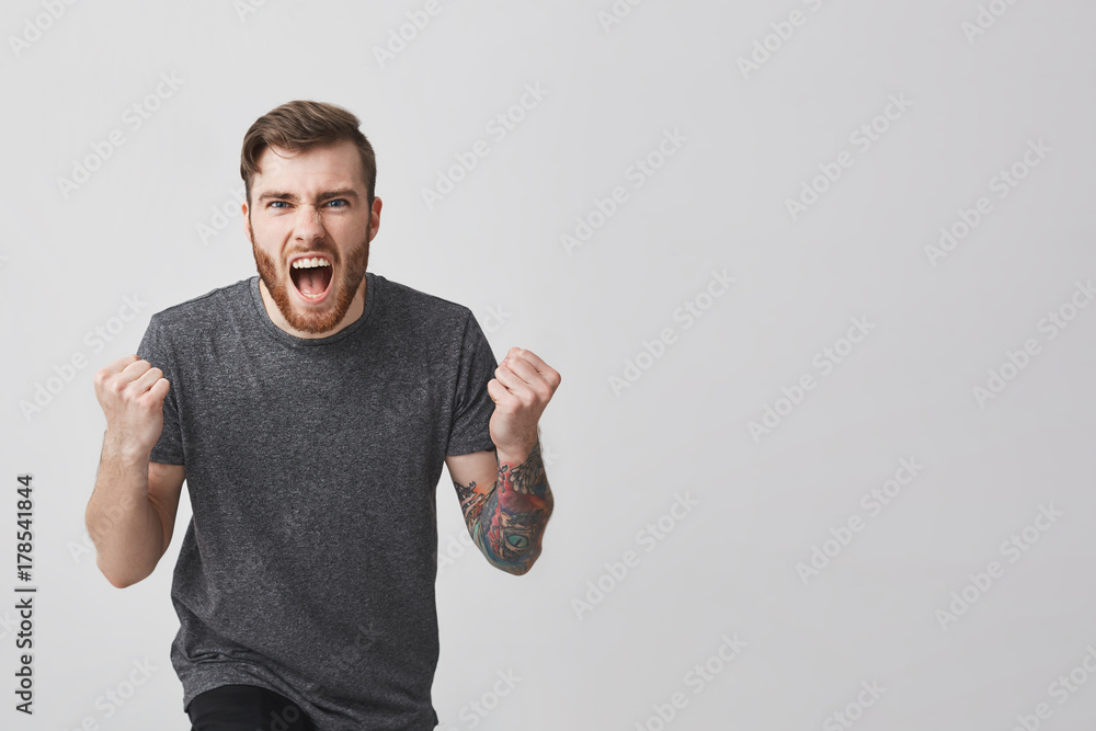 Portrait of young handsome bearded european hipster guy with tatooed arm and trendy haircut in casual stylish gray shirt holding fists in front of him, screaming loudly cheering for favourite hockey Portrait of young handsome bearded european hipster guy with tatooed arm and trendy haircut in casual stylish gray shirt holding fists in front of him, screaming loudly cheering for favourite hockey
