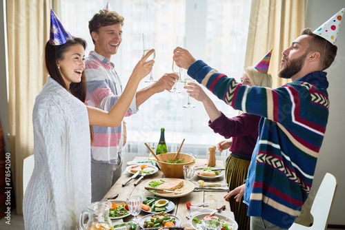 Group of happy young people wearing holiday caps celebrating Birthday with friends clinking champagne glasses and toasting during dinner party at home