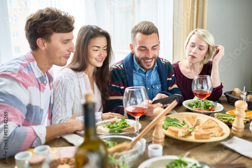 Portrait of four young people smiling happily watching videos from smartphone at dinner table during holiday celebration