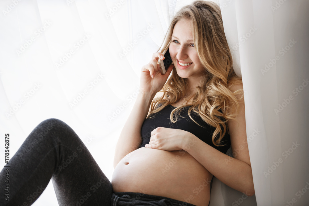 Close up of young good-looking cheerful future mother with long light hair in black clothes sitting on window sill in bedroom, happily talking with husband about pregnancy. Close up of young good-looking cheerful future mother with long light hair in black clothes sitting on window sill in bedroom, happily talking with husband about pregnancy.