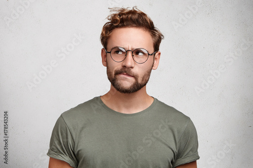 Serious thoughtful clever scientific worker wears spectacles, has beard, curves lower lip, tries to imagine new theory or thigs how to prove new axiom. Pensive unshaven man being very meticulous