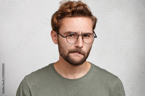 Suspicious bearded man looks doubtfully, curves lips, being indecisive, makes grimace, tries to find solution. Uncertain upset pensive male hesitates what to buy. Facial expressions concept.