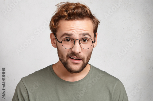 Clever handsome man has indignant expression, looks in bewilderment, tries to understand what he should do. Attractive bearded man curves lips as wants to say something. Facial expressions concept
