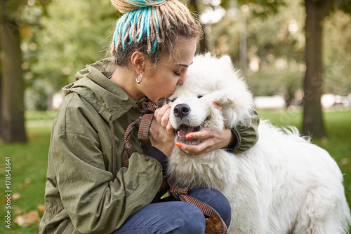 portrait of a young woman in a green coat and dreadlocks, kissing with his snow-white Samoyed dog in the autumn Park