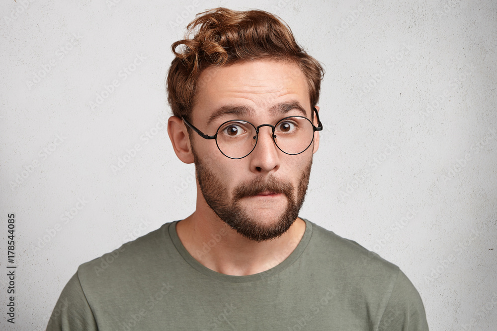 Pleasant looking bearded man presses lips, looks with astonishment, tries to understand something, being amazed or shocked. Indignant male adult has confused expression, poses against white wall Pleasant looking bearded man presses lips, looks with astonishment, tries to understand something, being amazed or shocked. Indignant male adult has confused expression, poses against white wall