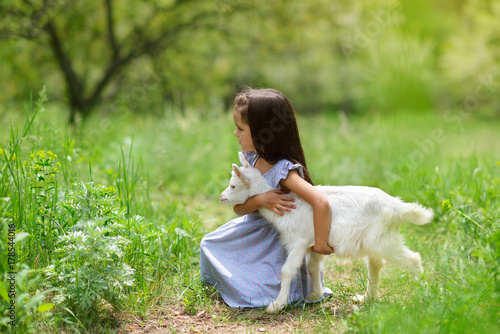 Little girl plays and huhs goatling in country, spring or summer nature outdoor. Cute kid with baby animal, countryside outdoor portrait, forest, trod. Friendship of child and yeanling photo