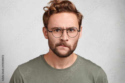 Puzzled serious irritated angry young unshaven man with beard and mustache curves lips, has sullen expression, being annoyed with girlfriend who betrayed him, sorts out relationships. Emotions concept