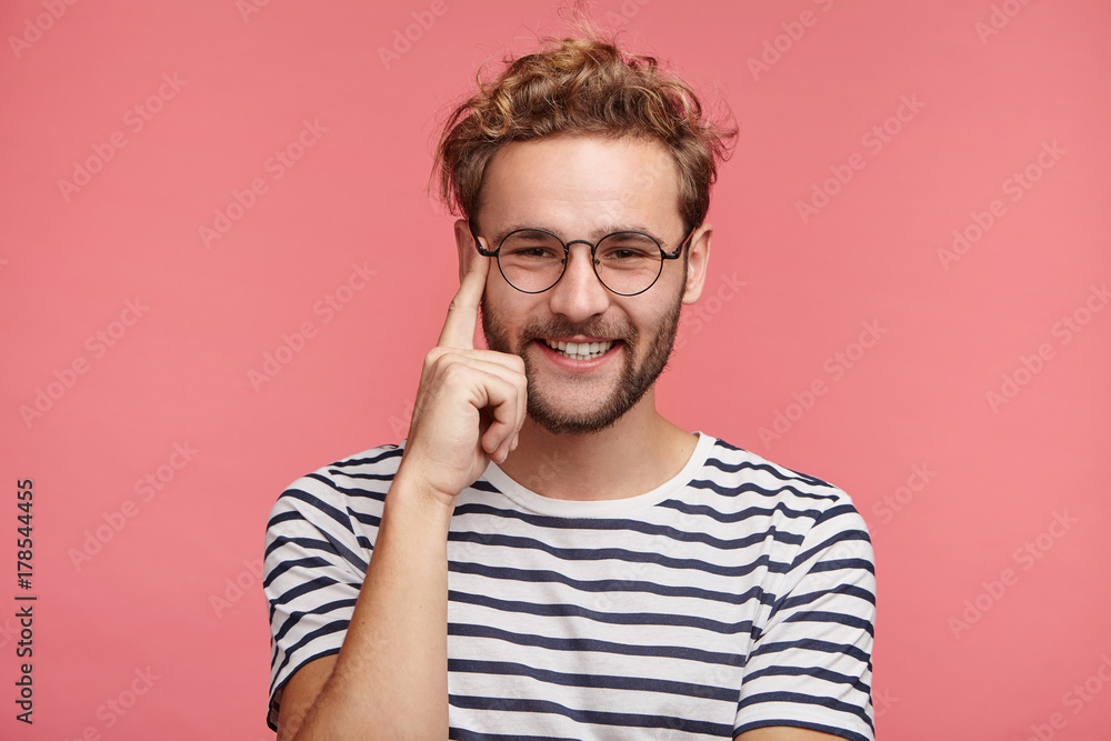Cheerful bearded young guy with confident expression keeps finger on temple, has gentle smile, rejoice good pleasant compliment from beautiful girl, isolated over pink background. Joy concept Cheerful bearded young guy with confident expression keeps finger on temple, has gentle smile, rejoice good pleasant compliment from beautiful girl, isolated over pink background. Joy concept