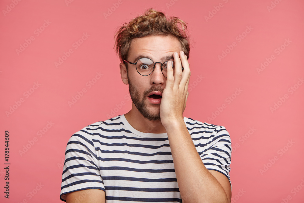Horrified bearded young man covers face with hand, being afraid to see awful scene in horror film, looks with opened mouth, amazed by heroes and actions. Emotional hipster scared of something Horrified bearded young man covers face with hand, being afraid to see awful scene in horror film, looks with opened mouth, amazed by heroes and actions. Emotional hipster scared of something
