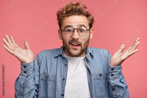 Waist up portrait of confused young man with trendy hairdo and stubble wears demin shirt and round spectacles, shrugs shoulders in bewilderment as makes difficult choice or decision in his life