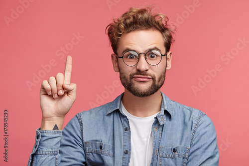 Funny fashionable man raises fore finger as gets idea, going to speak his mind, isolated over pink background. Male with specific appearance gestures in studio alone, has awkward expression.