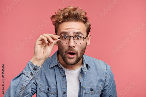 Terrified man dressed casually, stares through glasses, being surprised find out about his success. Male doesn`t believe his own luck, isolated over pink background. Emotions, people concept photo