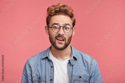 Unhappy handsome bearded young male frowns face in displeasure, looks directly into camera, wears spectacles and denim shirt, expresses hesitation or doubt as being asked disputable question
