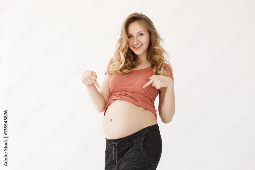 Portrait od joyful happy mama with blond hair in red t-shirt smiling brightfully, pointing on her belly with hands, posing for husband who wants take photo of pregnant wife. Portrait od joyful happy mama with blond hair in red t-shirt smiling brightfully, pointing on her belly with hands, posing for husband who wants take photo of pregnant wife.