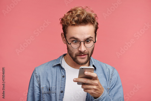 Shocked stylish hipster guy reads news online, uses free internet connection on modern cell phone, finds out about car accident in neigbour strict. Astonished male model recieves strange message photo