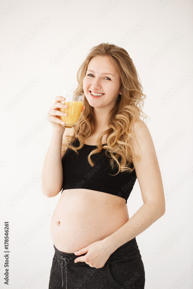 Vertical portrait of happy joyful european pregnant woman with blond curly hair in home clothes with opened belly, smiling brightfully, holding glass of orange juice going to take a sip. Vertical portrait of happy joyful european pregnant woman with blond curly hair in home clothes with opened belly, smiling brightfully, holding glass of orange juice going to take a sip.