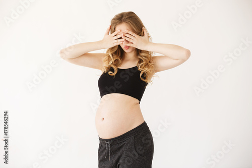 Portrait of funny attractive european mother with blond curly hair in black sport suit with opened pregnant belly  clothing eyes with hands  having surprised expression. Pregnant woman playing with
