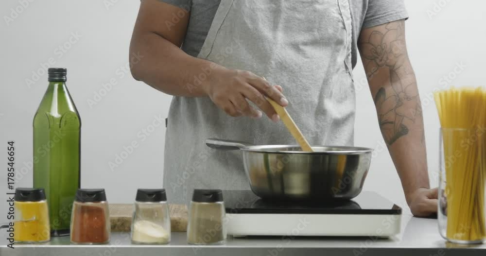 Attractive man in an apron mixes something in a stainless steel pan on heat with spices and oil around Attractive man in an apron mixes something in a stainless steel pan on heat with spices and oil around