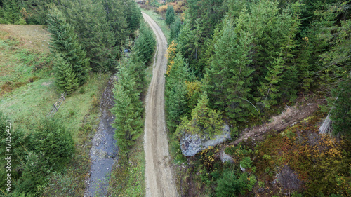 Fototapeta Naklejka Na Ścianę i Meble -  Aerial  view of a dirt road passing between a rock and a mountain river in spruce forest.
