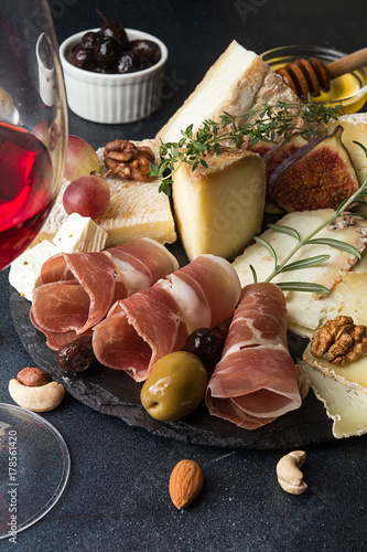 Glass of red wine and cheese plate with pieces moldy cheese, prosciutto, pickled plums, olives, grapes, figs, honey, pear, nuts on black slate photo