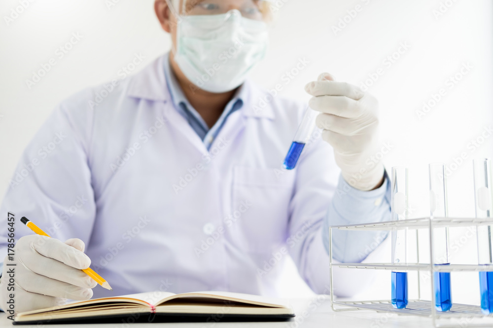 Scientist in white coat holding and examining test tube with reagent making notes of his research in laboratory Scientist in white coat holding and examining test tube with reagent making notes of his research in laboratory