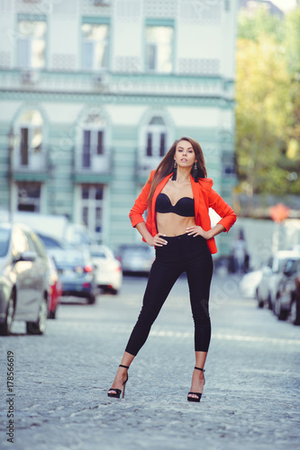 Fashionable look, hot day model of a young woman walking in the city, wearing a red jacket and black pants, blond hair outdoors over the city warm background