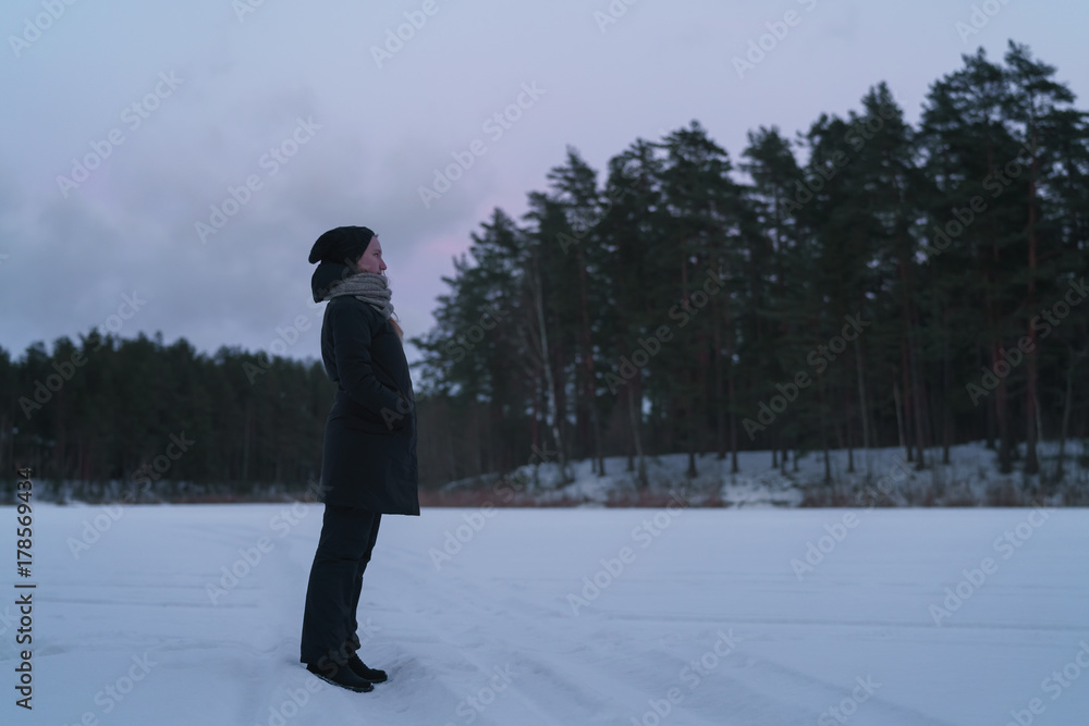 teenage girl standing on frozen lake in forest at evening in winter