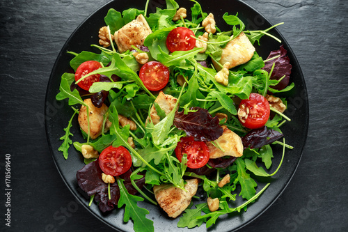 Fresh salad with chicken breast, arugula, nuts and tomatoes on black plate in a wooden table.