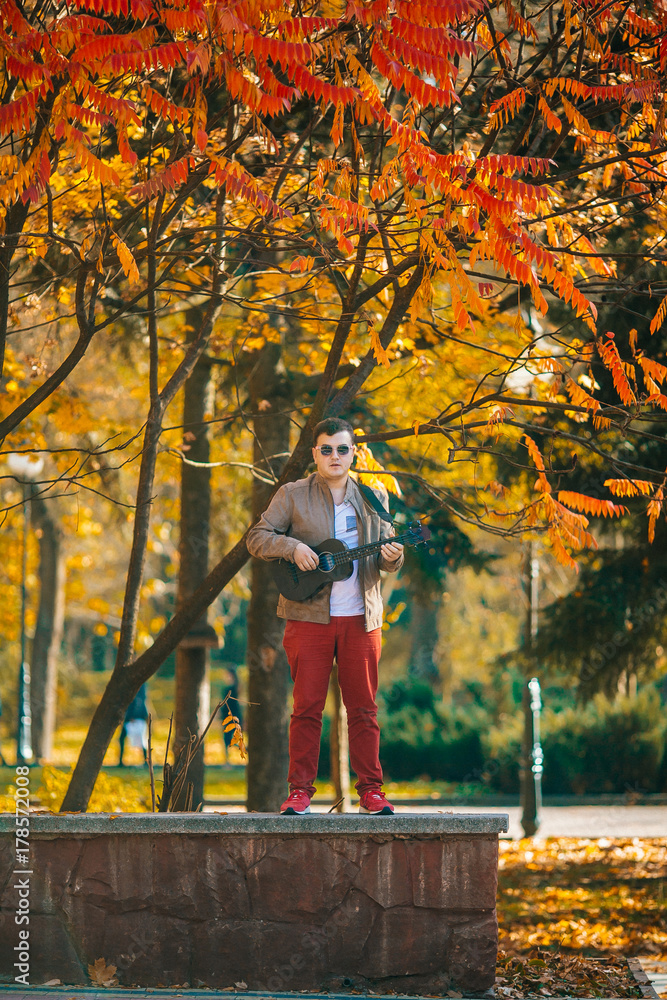 The young musician plays bass ukulele on the street. Man sitting on the ground with guitar. Concept. Basking The young musician plays bass ukulele on the street. Man sitting on the ground with guitar. Concept. Basking