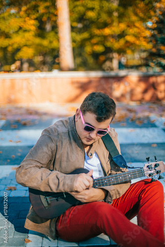 The young musician plays bass ukulele on the street. Man sitting on the ground with guitar. Concept. Basking