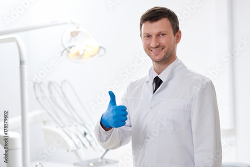 Cheerful mature dentist smiling happily showing thumbs up standing at his dental clinic dentistry healthcare positivity achievement success gesture maturity doctor practitioner.