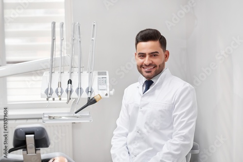 Young cheerful male professional dentist smiling to the camera joyfully sitting at his office copyspace dental oral examination checkup professionalism occupation medicine doctor.