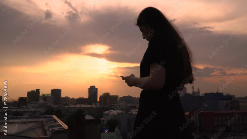 The girl uses phone on a roof. The girl looks in phone against the background of the centre of city.. The girl uses phone on a roof. The girl looks in phone against the background of the centre of city..