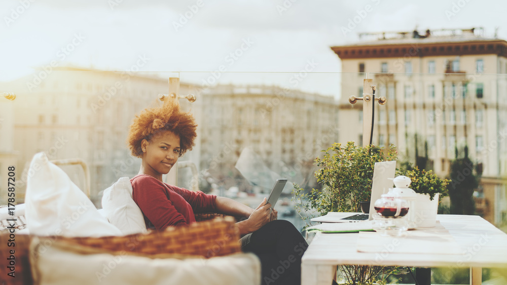 Charming young naughty black woman sitting on the wicker bengh outdoors in  roof restaurant with digital tablet; biracial teenage female freelancer in  street cafe with digital pad, laptop, and teapot Stock Photo