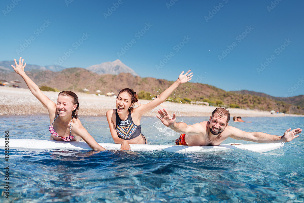 A multinational group of three friends of students laugh and swim in the sea with a board near the beach. Concept of travel and friendship A multinational group of three friends of students laugh and swim in the sea with a board near the beach. Concept of travel and friendship