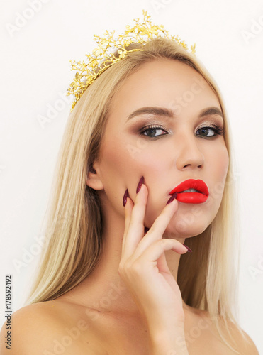 Beautiful stylish blonde girl with long wavy hair with a Golden crown on his head. Young Woman face portrait. Model with bright eyebrows, perfect make-up, red lips, touching her face. Sexy lady makeup
