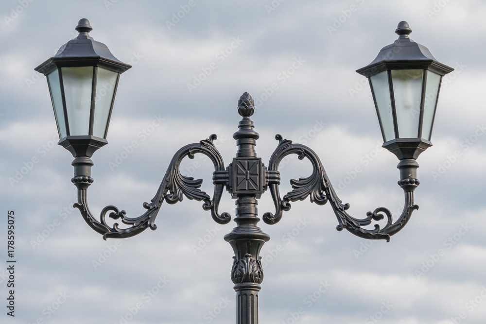 Two lamps on a wrought metal lamppost in retro style on a background of cloudy sky