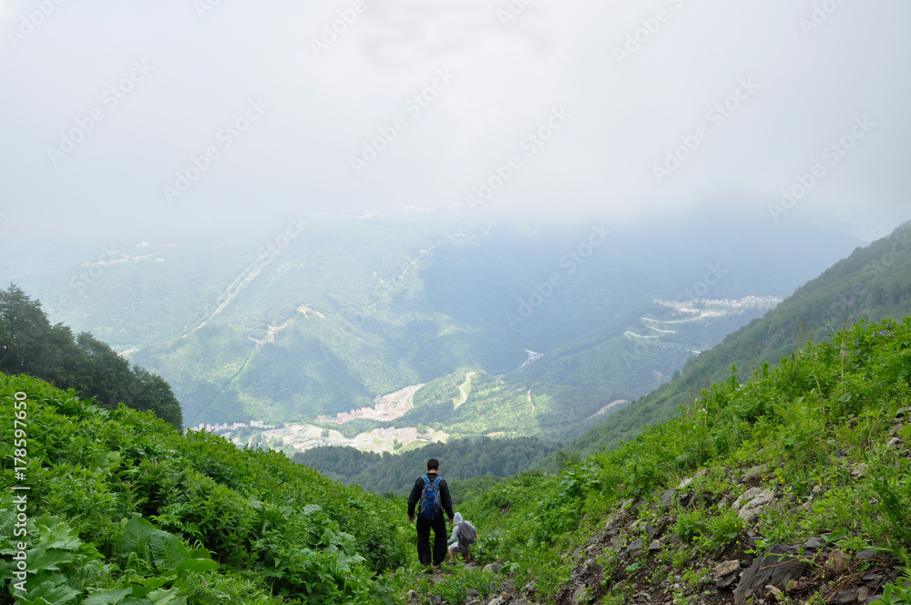 Two young men, come back from a campaign, climbing down a mountain among green vegetation and against the background of clouds and the town is below.