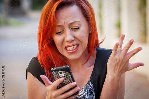 Φωτογραφία Cute Caucasian girl with red hair holding cellular phone in her hands with an upset, irritated look