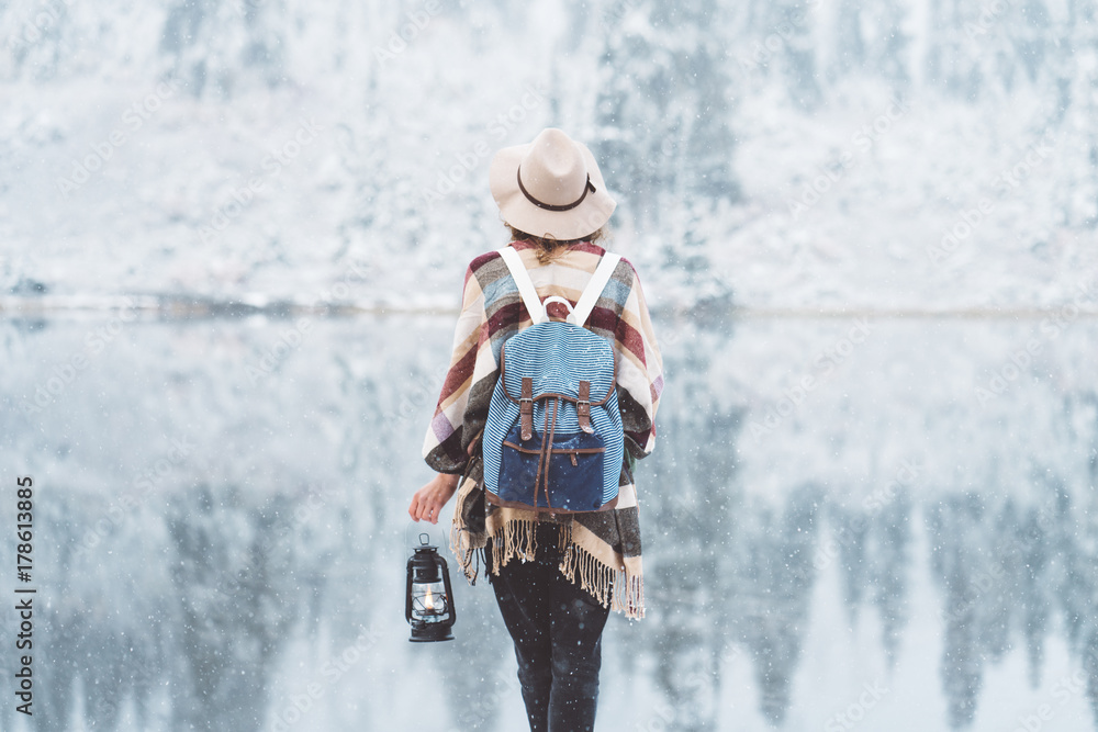 Traveling woman standing in the wild in front of amazing lake view with oil lamp. Wearing hat, poncho and backpack. Winter is coming, first snowfall. Wanderlust and boho style