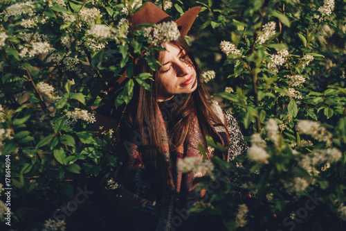 beautiful hipster girl in hat posing in green leaves with flowers in evening sunlight among shadows. stylish woman traveler calm portrait in spring garden. space for text. atmospheric moment